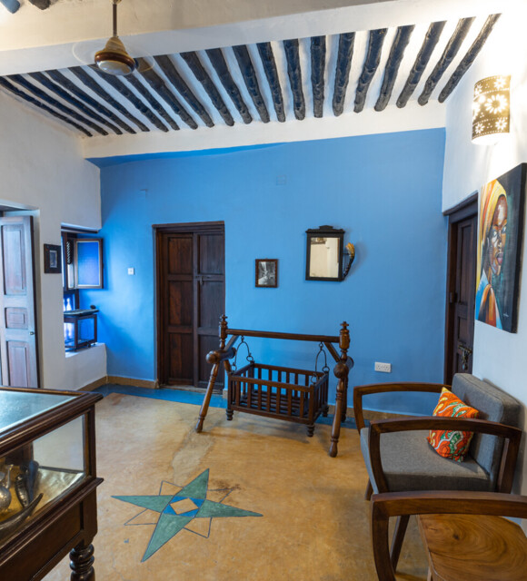 Cozy lounge with blue accent wall, traditional wooden cradle, vintage ceiling beams, carved wooden furniture, and colorful cushions