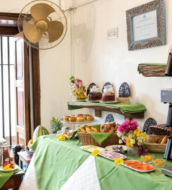 Bright breakfast buffet with pastries, muffins, cereal jars, fresh fruit, coffee machine, and condiments on green tablecloth in rustic dining area
