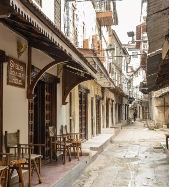 Narrow street with Shaba Boutique Hotel featuring wooden chairs and tables outside, traditional architecture, and weathered buildings