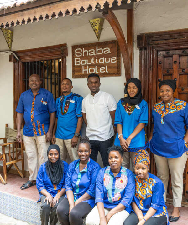 Hotel staff in vibrant blue and patterned uniforms pose outside Shaba Boutique Hotel's entrance with wooden doors and traditional décor