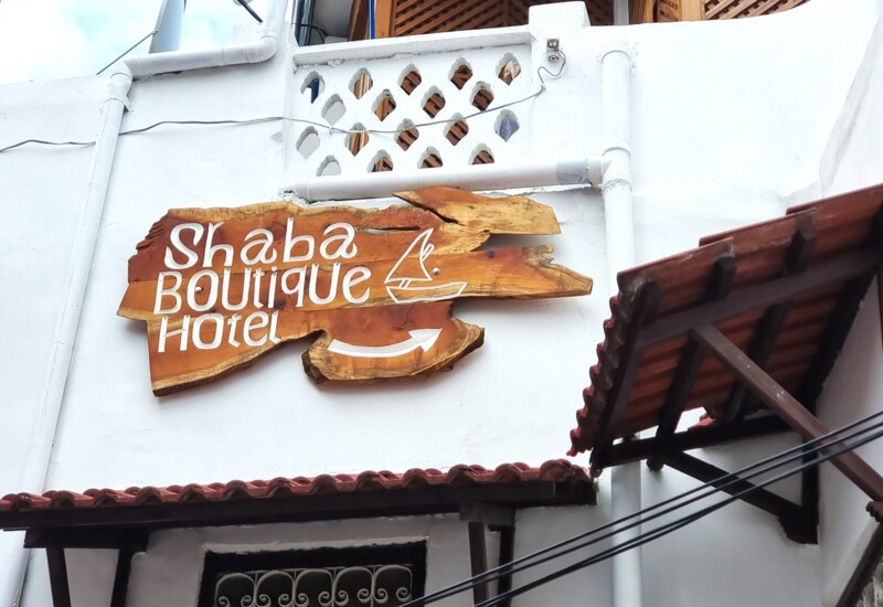 Wooden sign for Shaba Boutique Hotel on white stucco wall with ornate ventilation blocks and terracotta roof awning