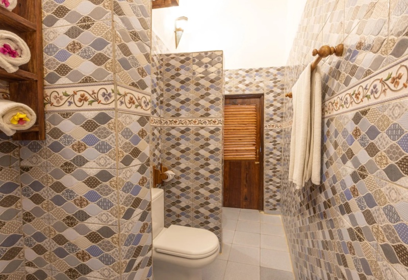 Hotel bathroom with colorful patterned tiles, wooden door, neatly rolled towels with flower accents, and wall-mounted towel rack