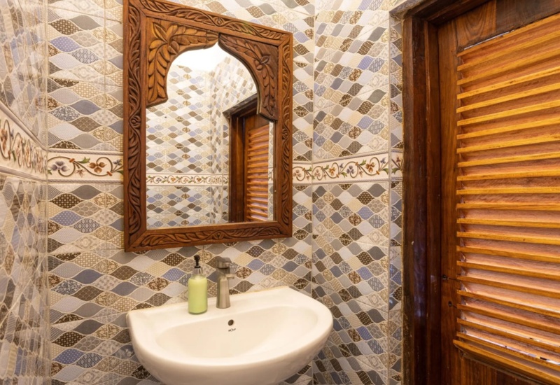 Compact bathroom with patterned tile walls, carved wooden mirror frame, white sink, and wooden louvered door.