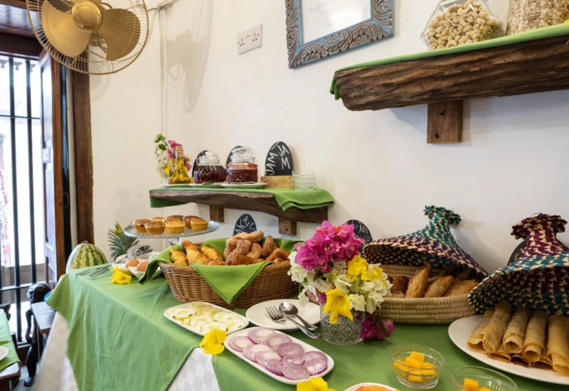 Breakfast buffet with assorted pastries, sliced onions, mango cubes, jam jars, and colorful fresh flowers on green tablecloth