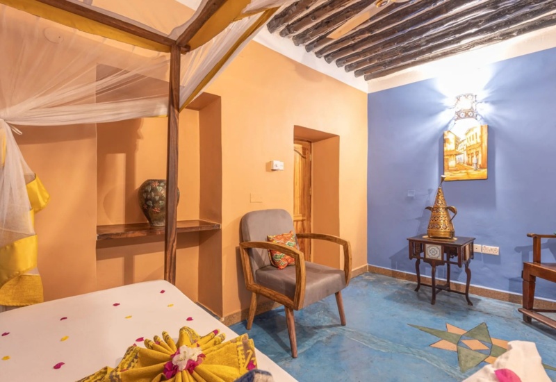 Colorful hotel room with wooden beamed ceiling, canopy bed with mosquito net, two wooden chairs with patterned cushions, and decorative vase
