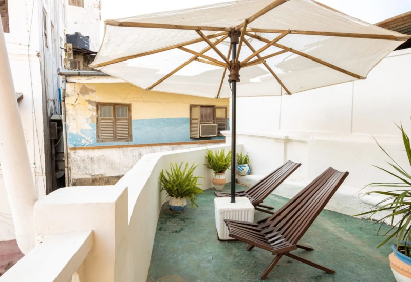 Cozy balcony with two wooden lounge chairs under a large umbrella, surrounded by potted plants and white walls