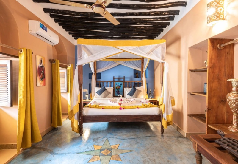 Bright room with wooden four-poster bed draped in white and yellow, decorative floor tile, rustic wooden shelves, and yellow curtains