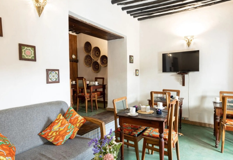 Cozy dining area with wooden tables and chairs, colorful cushions, wall decor, and a mounted flat-screen TV in a bright, rustic setting