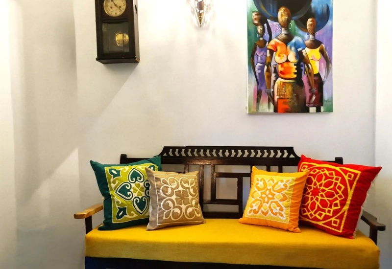 Cozy corner with a wooden bench featuring a yellow cushion and four colorful patterned throw pillows beneath wall art and a vintage clock.