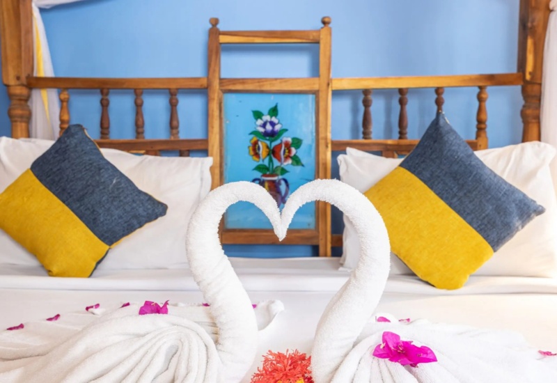 Romantic hotel room with wooden four-poster bed, blue wall, colorful floral artwork, and towel swans decorated with pink petals.