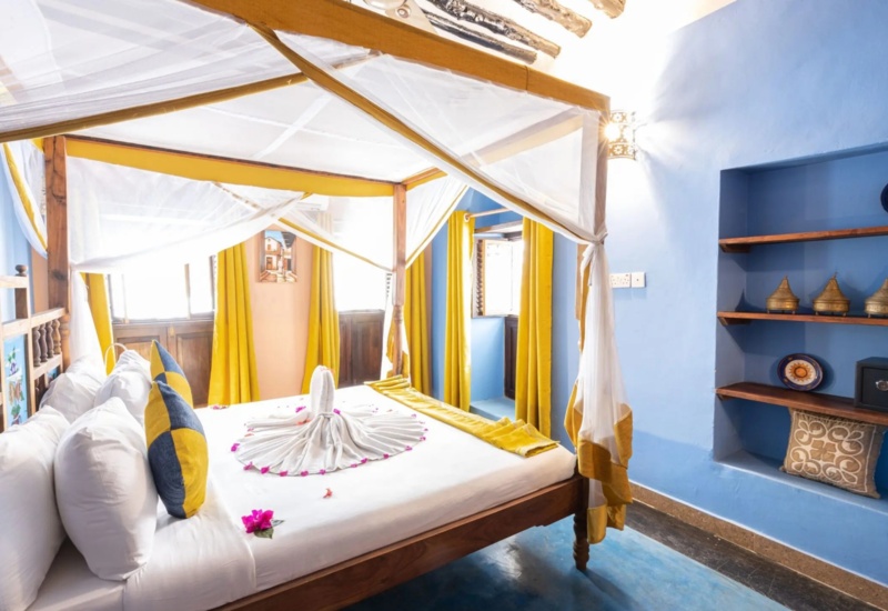 Bright room with wooden four-poster bed draped in white netting, blue walls, yellow curtains, and decorative shelves with local art and safe.