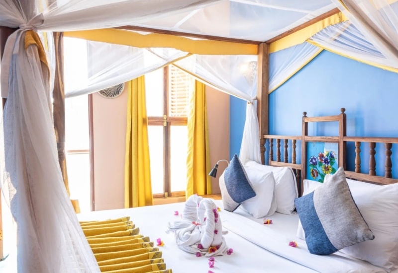 Bright hotel room with a four-poster bed featuring blue and white pillows, yellow curtains, and towel swans with flower petals