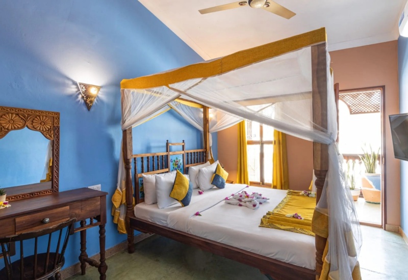 Bright hotel room with a wooden four-poster bed draped in yellow and white linens, a carved wooden desk, blue walls, and balcony access