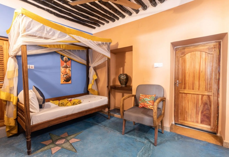 Cozy room with a wooden four-poster bed draped in yellow and white fabric, vibrant wall art, and a cushioned chair with colorful pillow