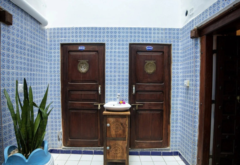 Traditional restroom area with separate wooden doors for men and women, blue patterned tiles, and a central hand-washing sink with floral decor