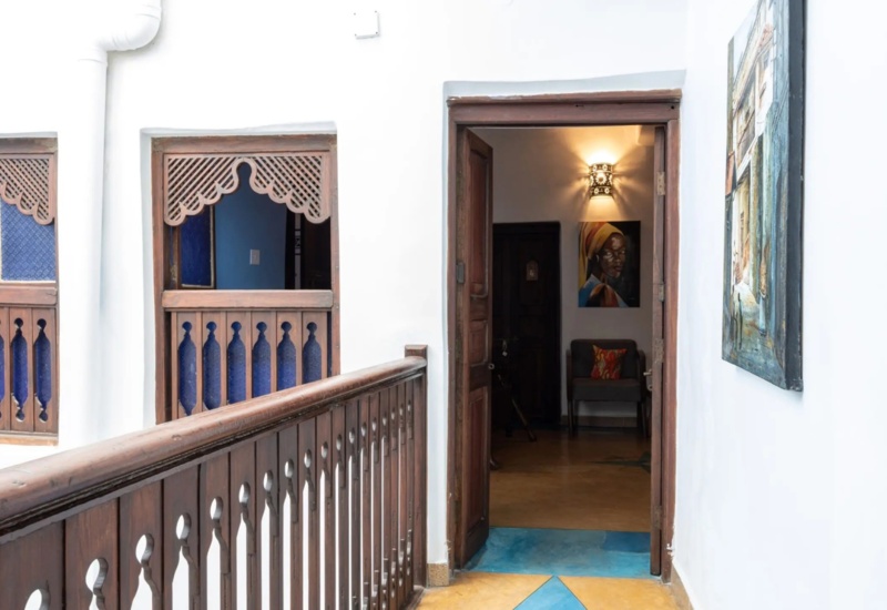 Hallway with wooden railing and carved windows leading to a room with warm light, artwork, and cozy seating with colorful cushions