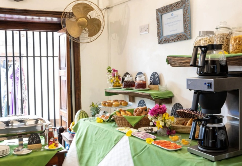 Breakfast buffet with fresh muffins, croissants, sliced fruits, jams, coffee, and cereals in a bright, airy dining area with rustic decor