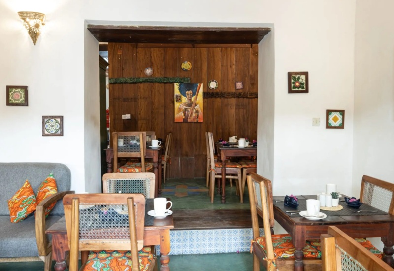 Cozy hotel dining area with wooden chairs and tables, colorful patterned cushions, artwork on white walls, and warm lighting