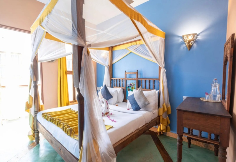 Cozy four-poster bed with white and yellow drapes in a bright room featuring blue accent wall and wooden furniture