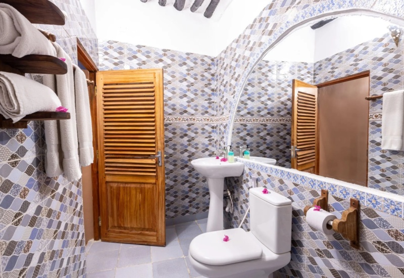 Bathroom with patterned blue and brown tiles, wooden doors, white towels on wooden racks, pedestal sink, and toilet with floral accents