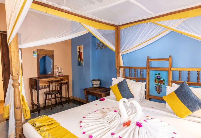Cozy hotel room with a wooden four-poster bed draped in white and yellow netting, decorative pillows, and towel swans on the bed.
