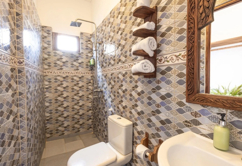 Bathroom with patterned tile walls, walk-in shower with rainfall showerhead, wooden towel rack, toilet, sink, and carved wood mirror frame
