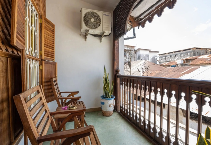 Cozy balcony with wooden chairs, decorative railing, potted plant, and views of rustic rooftops under a sunny sky