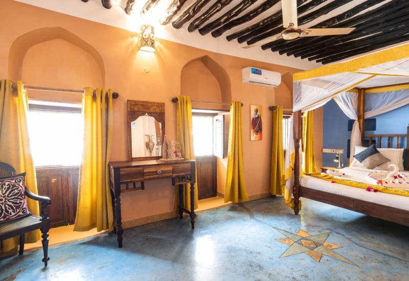 Cozy room with four-poster bed, traditional carved furniture, bright yellow curtains, blue accent wall, and decorative floor tile