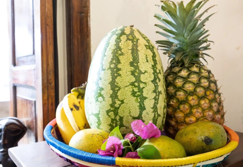 Colorful fruit basket with watermelon, pineapple, bananas, mangoes, and pink flowers on a wooden surface near rustic window and carved sculpture