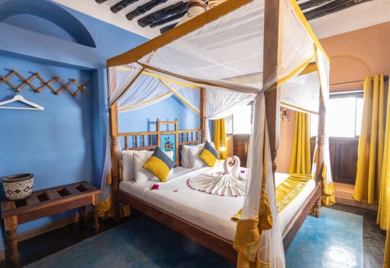 Cozy four-poster bed with white and yellow drapes, decorative pillows, and towel swans in a vibrant blue and yellow room with wooden accents