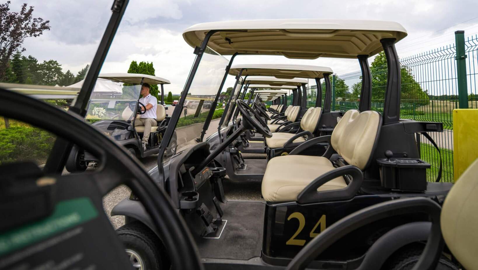 Rząd numerowanych wózków golfowych z beżowymi siedzeniami i daszkami, zaparkowanych przy ogrodzeniu; w tle osoba siedzi za kierownicą.