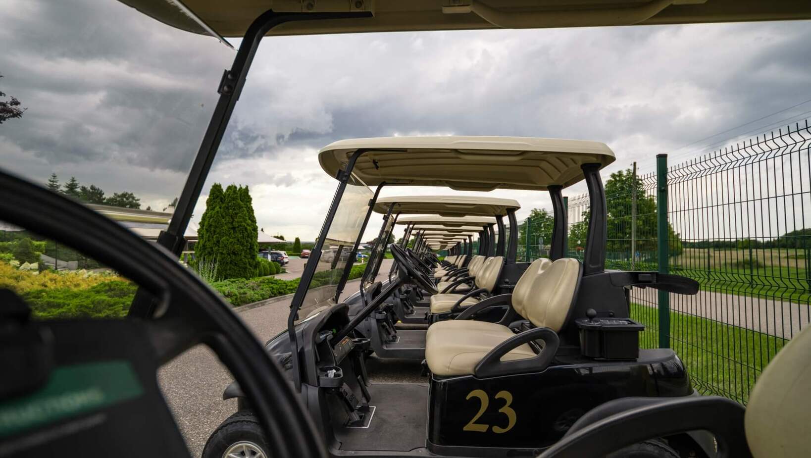 Rząd wózków golfowych z beżowymi siedzeniami (nr 23), gotowych do wypożyczenia przy polu golfowym, pochmurny dzień.