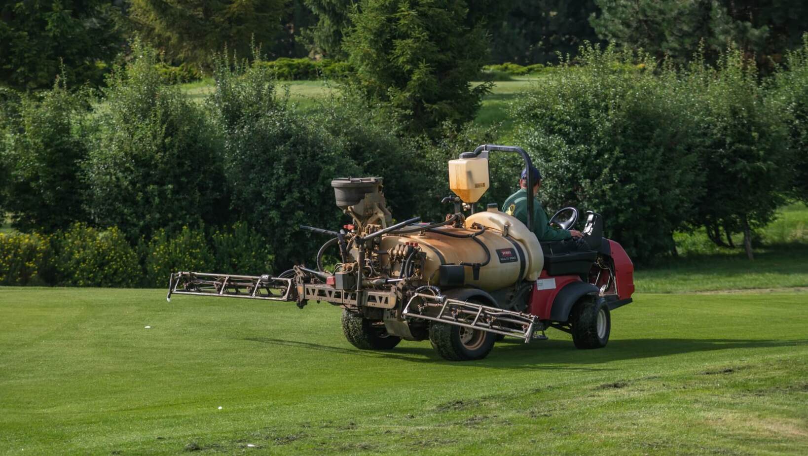 Pracownik obsługujący ciągnik z rozpylaczem pielęgnujący trawnik pola golfowego, otoczone drzewami.