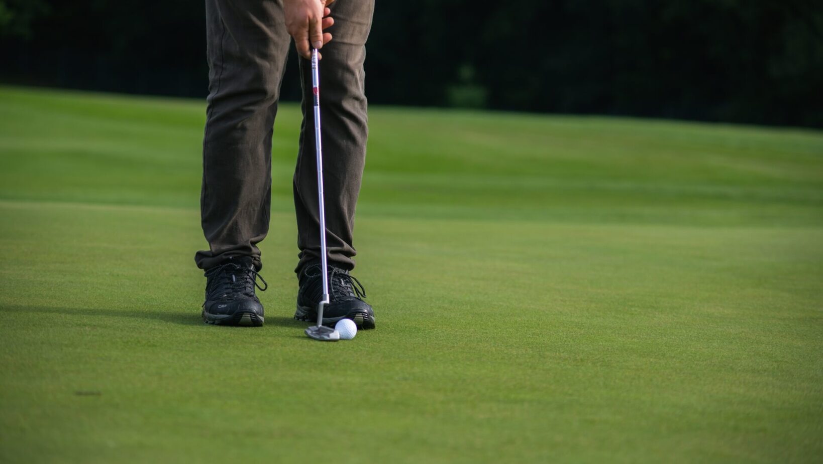 Gość na hotelowym polu golfowym wykonuje putt na gładkim greenie, widać putter, piłkę i sportowe buty