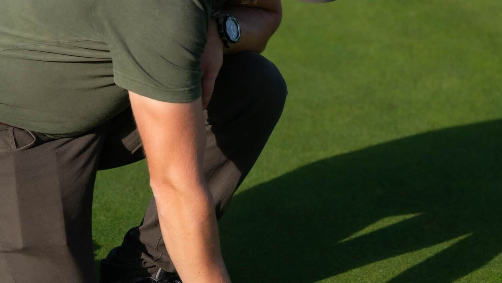 Pracownik hotelowego pola golfowego klęczy na idealnie przyciętym greenie, używa metalowego narzędzia do przygotowania dołka.