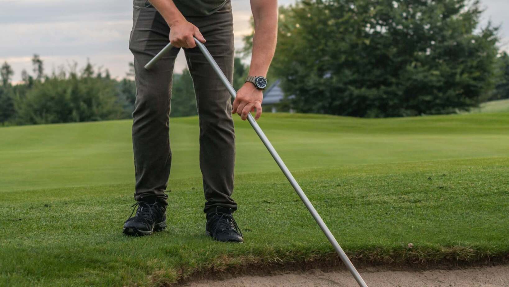 Obsługa pola golfowego grabiąca bunkier przy zielonym trawniku, przygotowując piasek na gości; spokojna, zachmurzona atmosfera