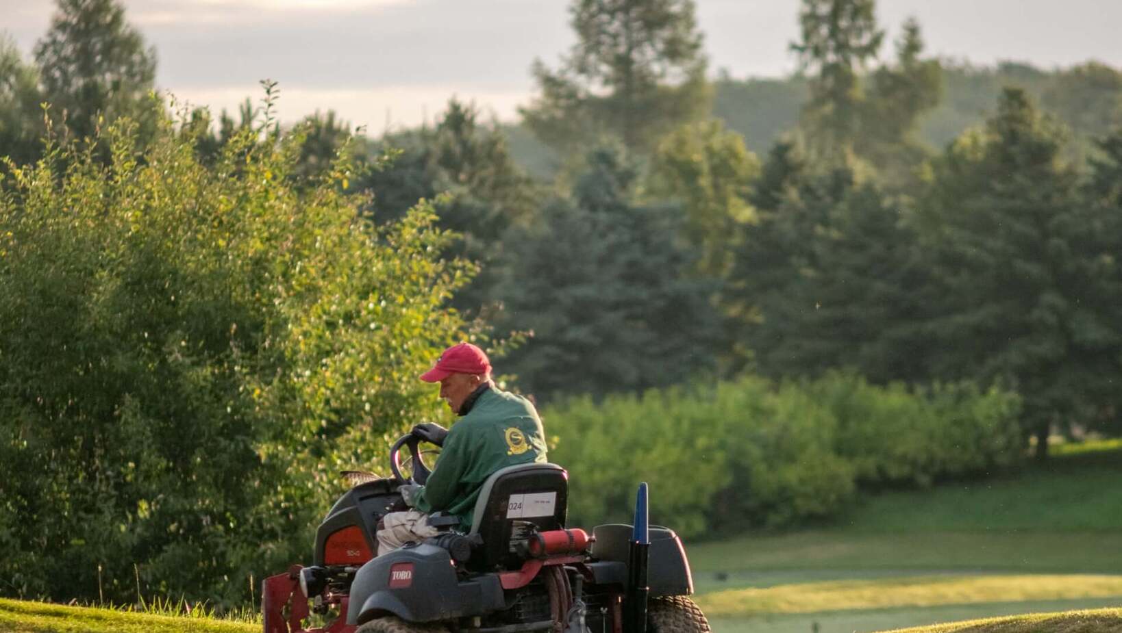 Pracownik obsługi trawników na traktorku pielęgnacyjnym wygładza bunkier na zielonym polu golfowym, poranne światło.