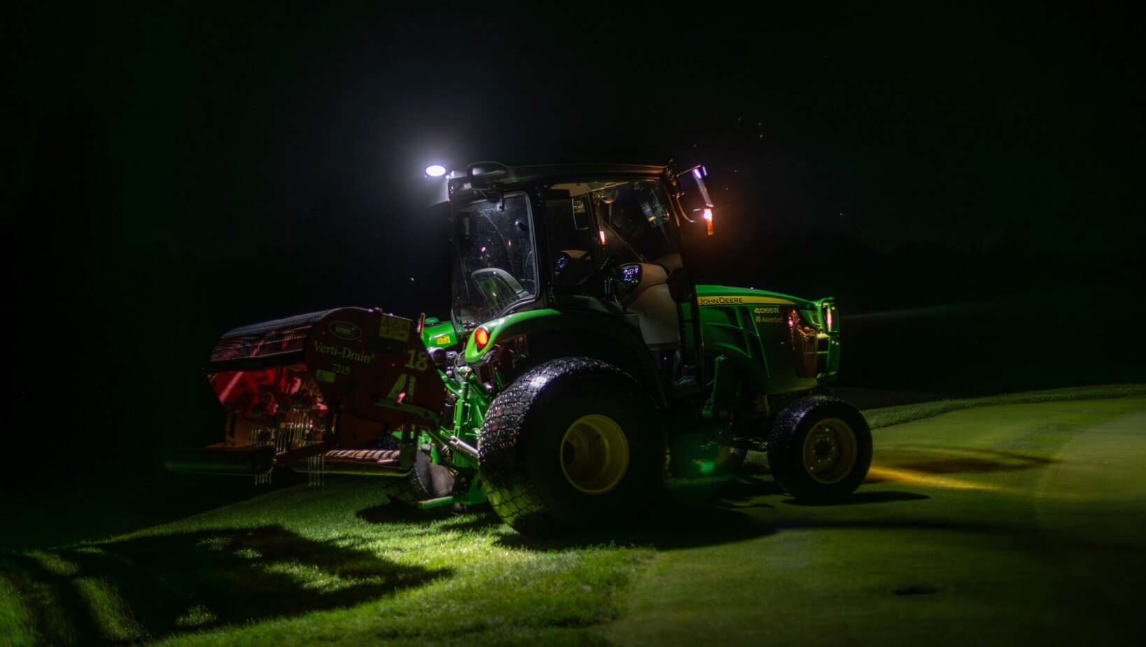 Traktor serwisowy na polu golfowym nocą, oświetlony reflektorami podczas aeracji trawnika.