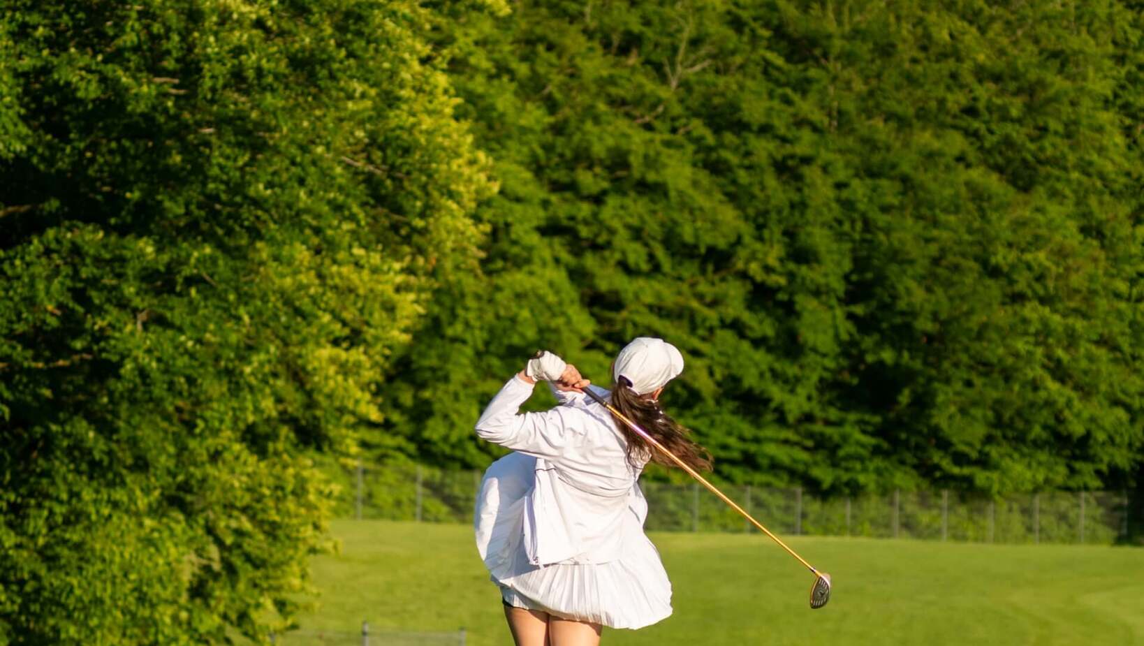 Golfistka w białej stylowej odzieży wykonuje zamach z tee na zielonym polu golfowym otoczonym drzewami