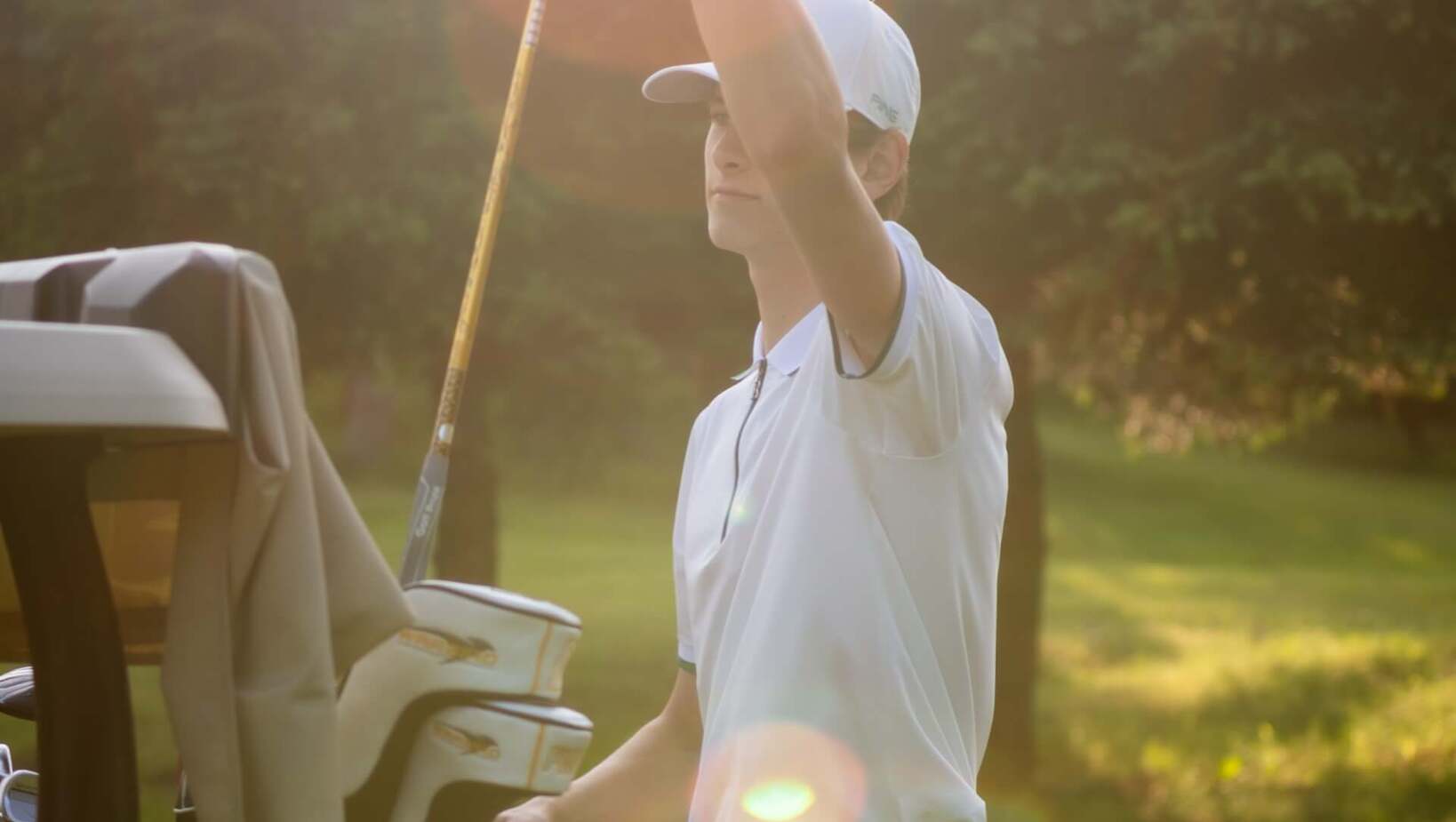 Gość w białej koszulce i czapce przy wózku golfowym podnosi kij na słonecznym polu, ciepła poświata i leśne tło