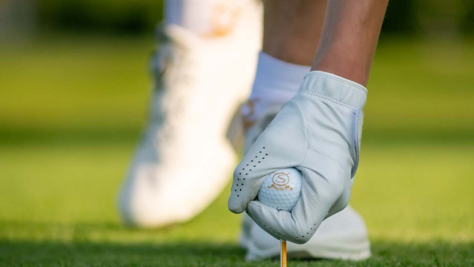 Ręka w białej rękawiczce golfowej ustawia piłkę na tee na zielonym polu golfowym, w tle białe buty