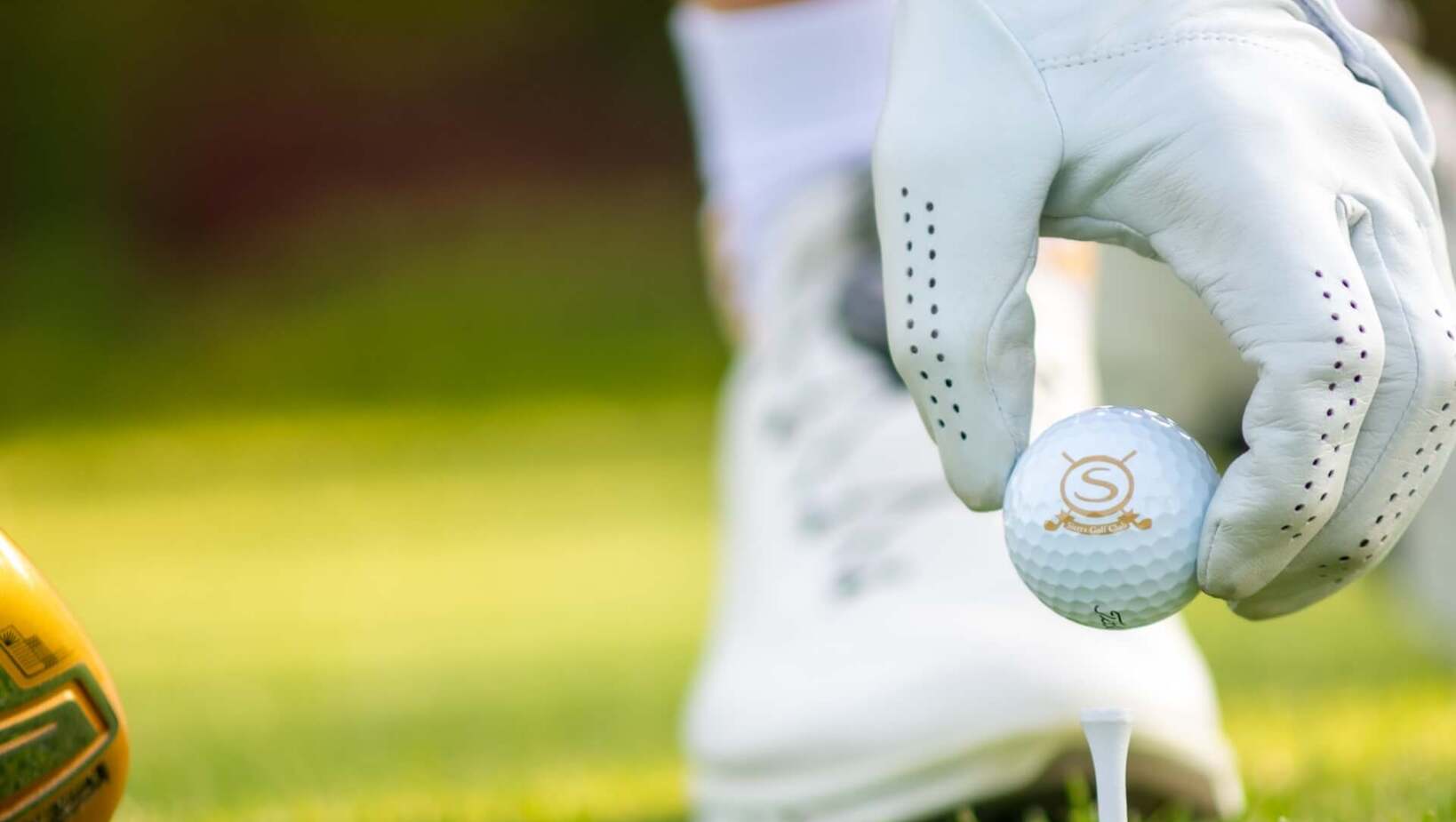 Ręka w białej rękawiczce ustawia piłkę na tee na zielonym polu golfowym resortu, w pobliżu kija i białego buta