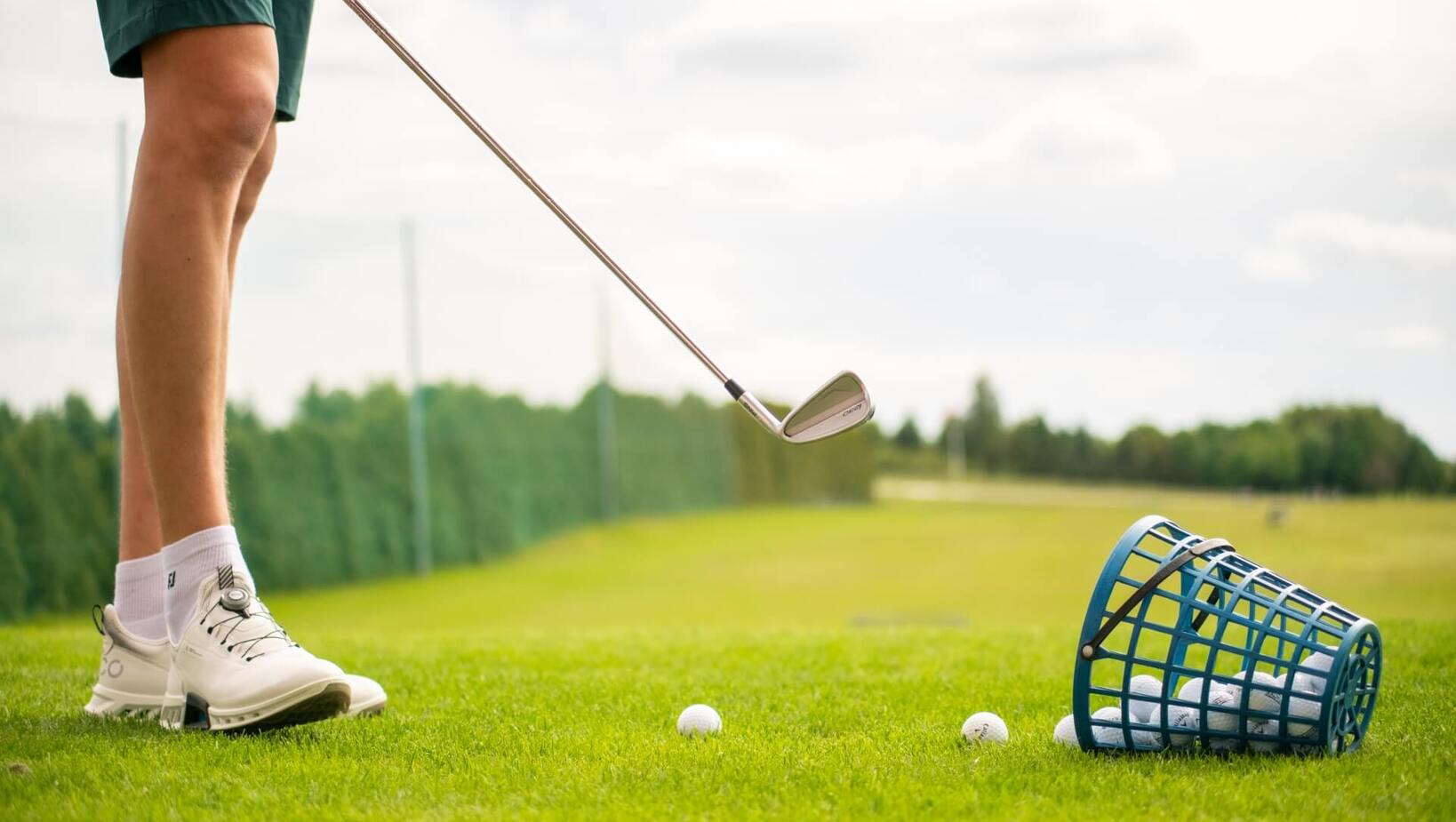 Nogi golfisty w białych butach, kij do golfa nad piłką i przewrócony kosz z piłkami na zielonym driving range.