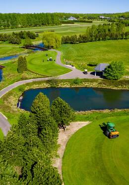 Rozległe pole golfowe z zadbanymi greenami, stawami i alejkami; widoczne wózki golfowe, altanka i traktor pielęgnacyjny.