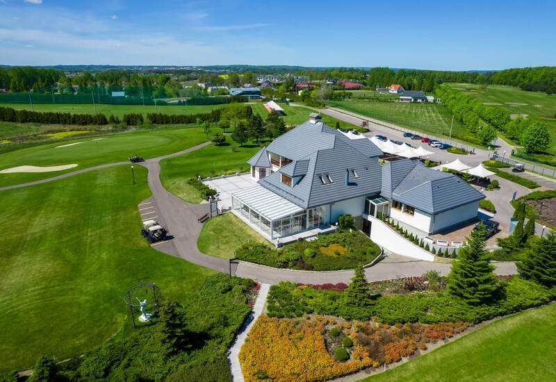 Klub golfowy z przeszkloną werandą, tarasem, miejscem parkingowym i wózkami golfowymi na zadbanych polach.
