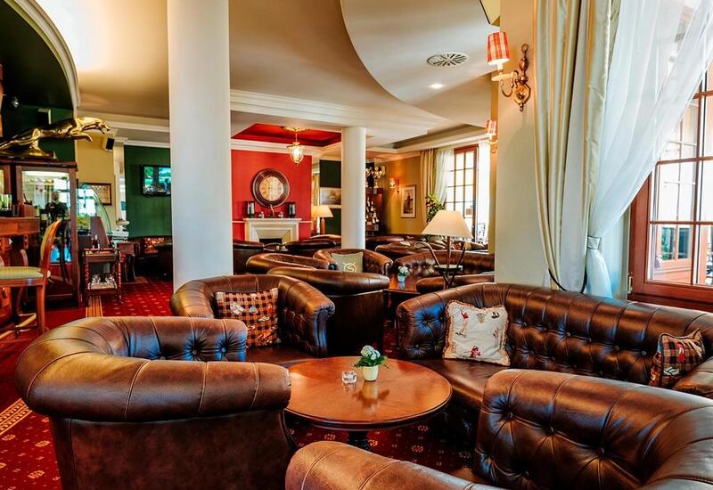 Przytulny hotelowy salon z brązowymi skórzanymi kanapami Chesterfield, okrągłymi stolikami, kominkiem, filarami i ciepłym oświetleniem.
