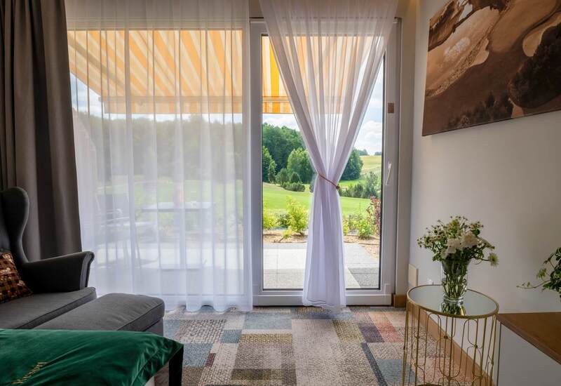 Salon apartamentu z przeszklonym wyjściem na taras, przewiązaną zasłoną, żółto-białą markizą i widokiem na zielone pole.