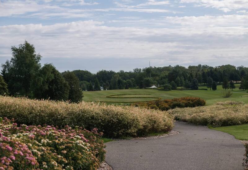 Zadbane tereny resortu z alejką, kwitnącymi krzewami i panoramicznym polem golfowym z ozdobnym okręgiem krzewów.