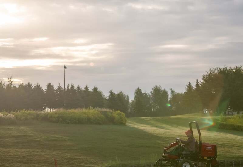 Wschód nad zadbanym polem golfowym, pracownik kosi trawę traktorkiem, równo przycięte fairwaye i rosa.