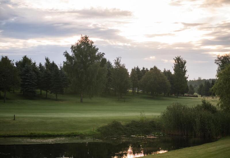 Pole golfowe z gładką trawą, stawem i rzędami drzew, spowite złotym światłem nieba